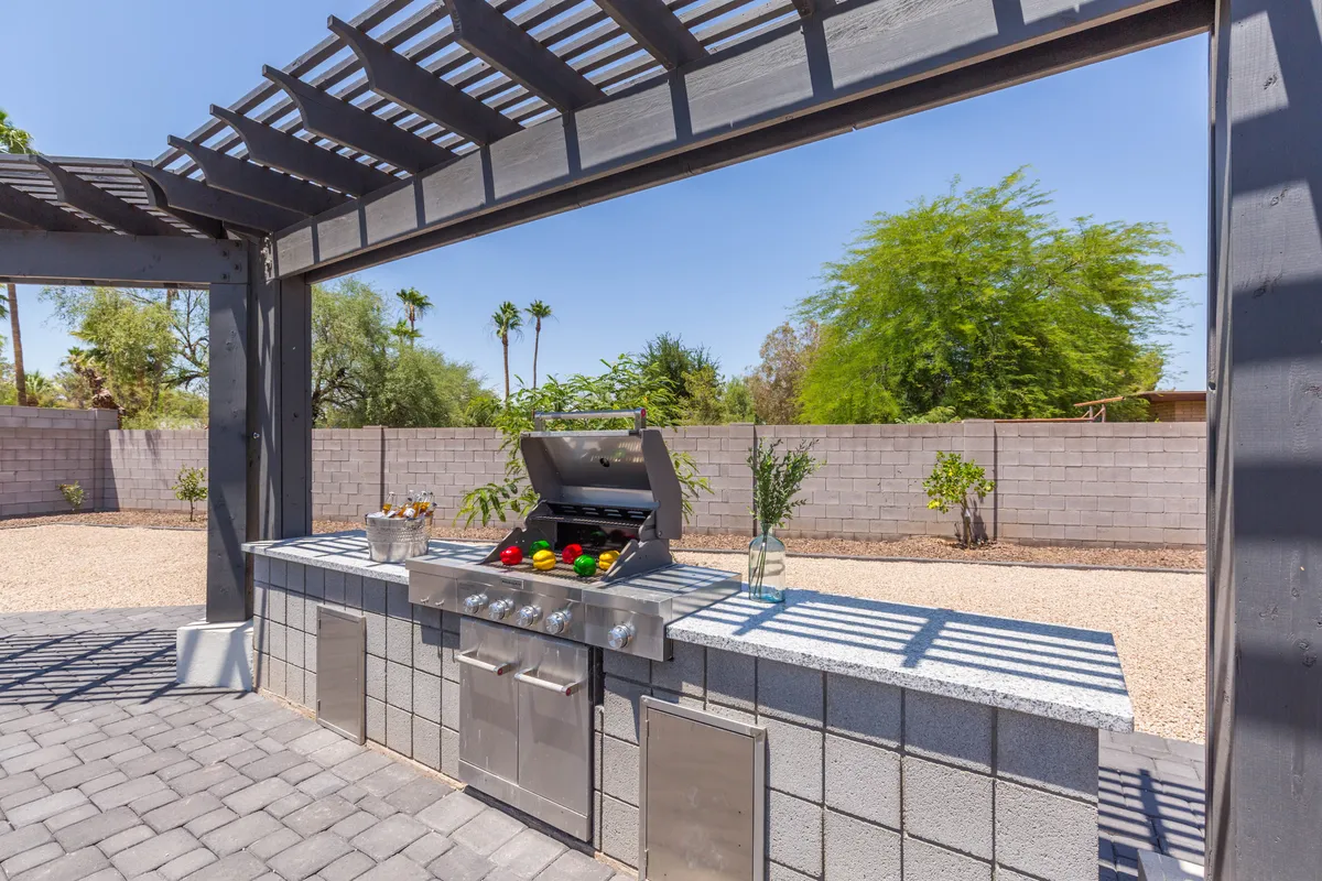 This image showcases an inviting outdoor grill area beneath a stylish pergola, surrounded by lush landscaping. The space is clearly designed for barbecuing, outdoor dining, and social gatherings in a relaxed, sunny setting.
