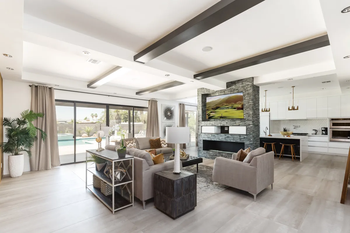 An expansive open-concept living area is showcased, with plush sofas surrounding a substantial coffee table, a large TV mounted on the wall, a kitchen island in the background, and wide windows flooding the space with natural light. The area is set up to encourage both leisurely conversations and engaging entertainment, with seamless flow between the living, dining, and kitchen spaces.