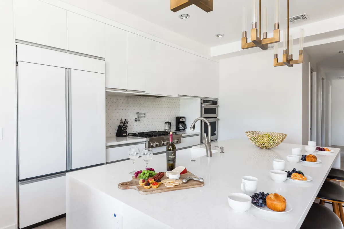 A modern kitchen featuring a gleaming white island set for entertaining, with wine glasses, a fruit platter, and chic pendant lighting overhead. Stainless steel appliances and stylish decor evoke a sense of both culinary sophistication and comfort, with ample space for casual gatherings or family breakfasts.