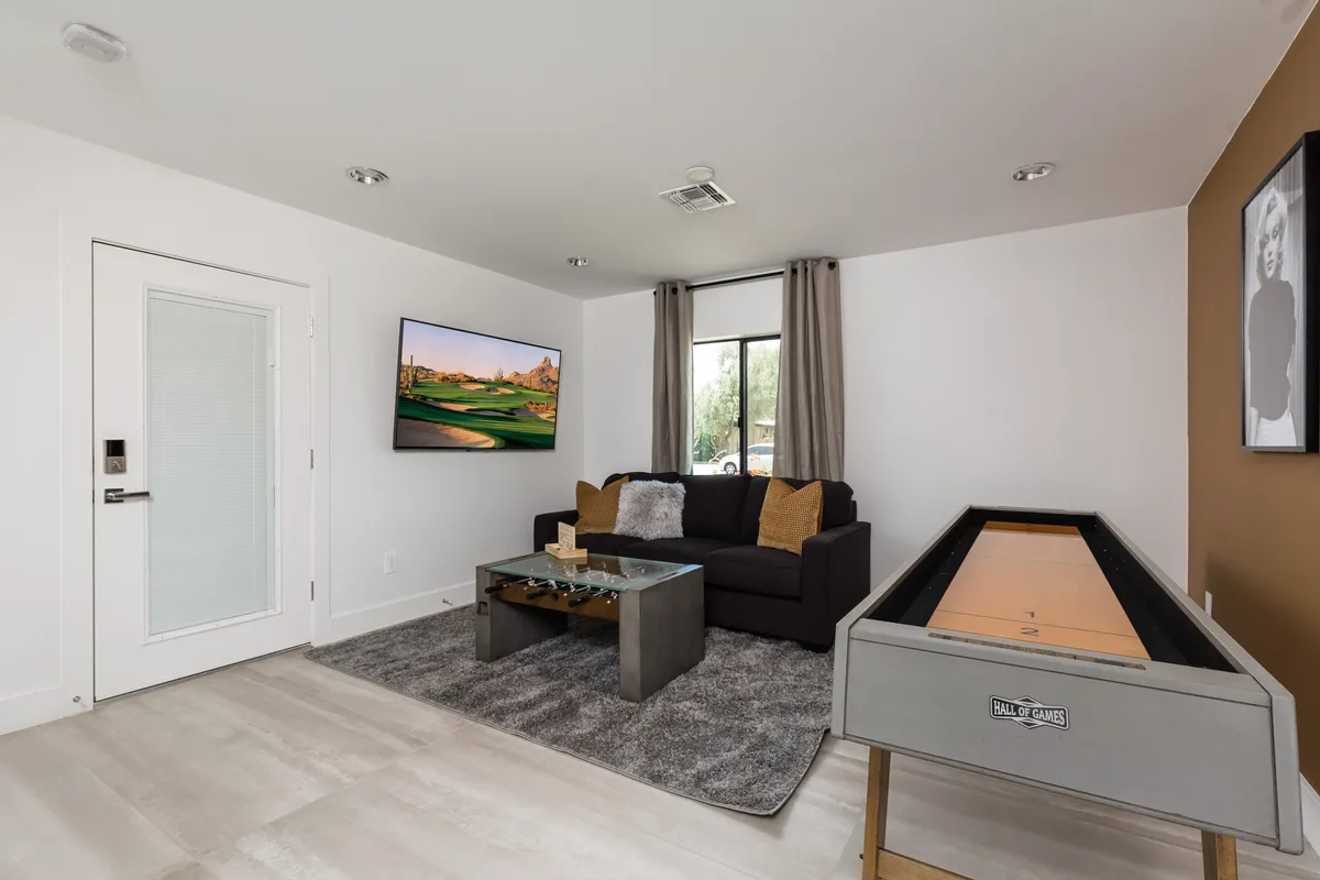 A comfortable interior game room featuring a shuffleboard table, sofa, television, and decorative artwork. The space is bright and casual, designed for game nights and relaxed socializing.