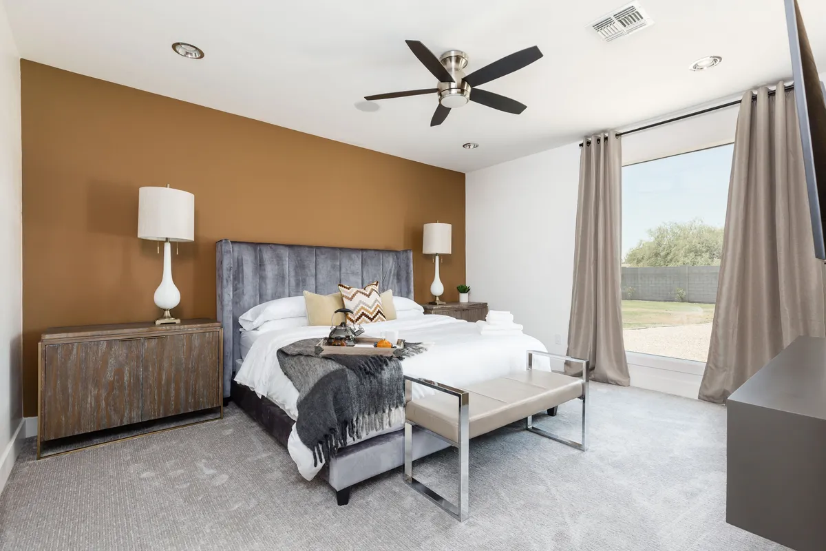 A serene and modern bedroom with a large bed, sunlit window, nightstand, and lamps, evoking a sense of peaceful comfort and inviting relaxation. The space is styled with tasteful decor and natural light, perfectly suited for restful evenings or quiet mornings.