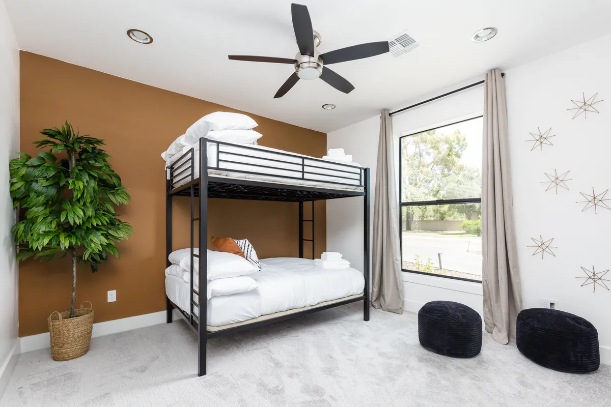 A playful and comfortable children’s bedroom featuring a modern bunk bed, decorative plant, large window, and black poufs, filled with natural light. The setup encourages both sleep and fun, ideal for family stays or group getaways.