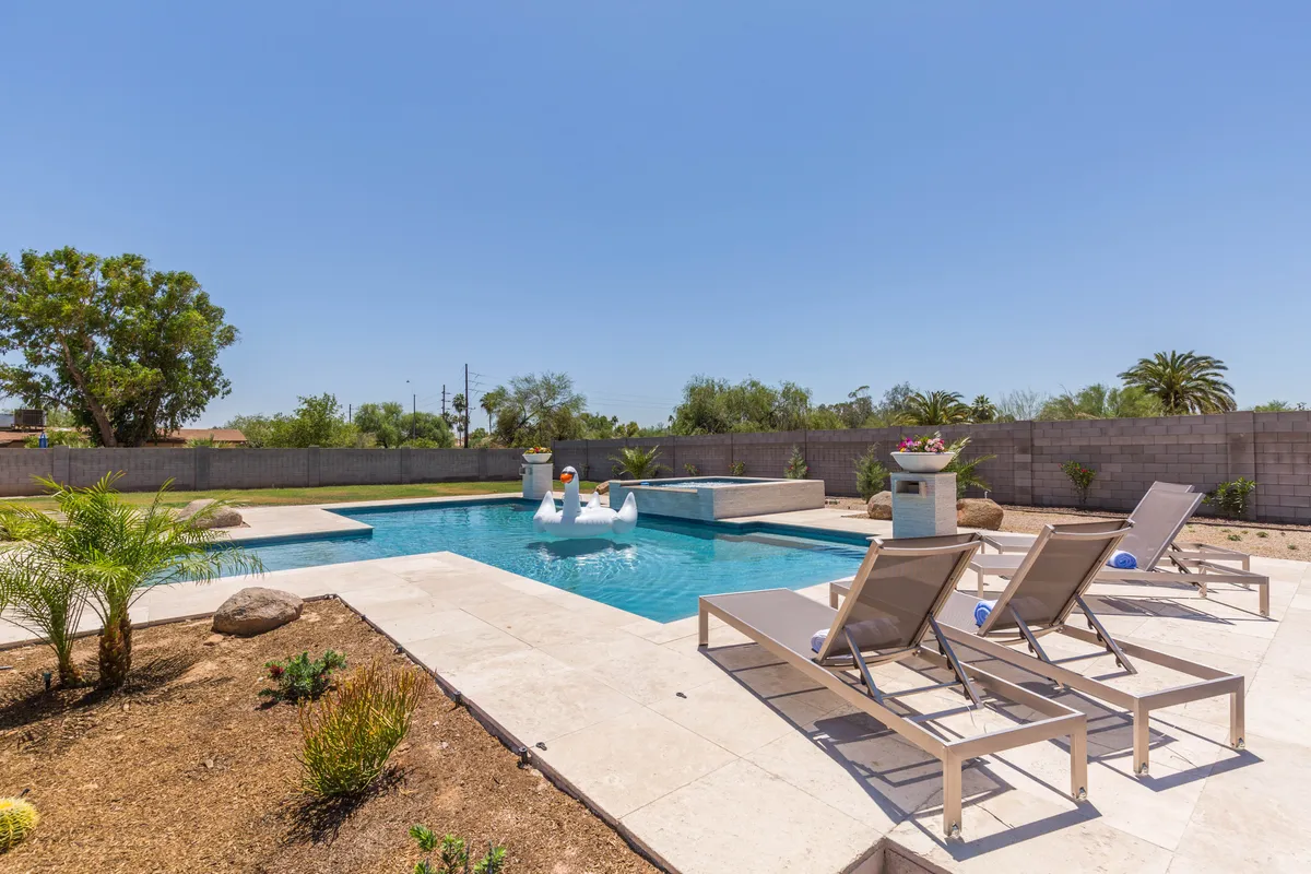 A chic backyard oasis featuring a sparkling swimming pool surrounded by comfortable lounge chairs, whimsical swan pool floats, and a landscaped garden. The atmosphere is tranquil and inviting, perfect for relaxation, poolside gatherings, or sunbathing in style.